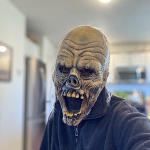 May include: A person wearing a scary, detailed, gray and brown zombie mask with a wide open mouth and visible teeth. The mask is worn over a black fleece jacket.