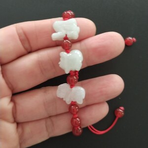 May include: A white jade elephant charm bracelet with red beads and a red cord. The bracelet features three elephant charms and is held in a hand against a dark background. The bracelet is a unique piece of jewelry.