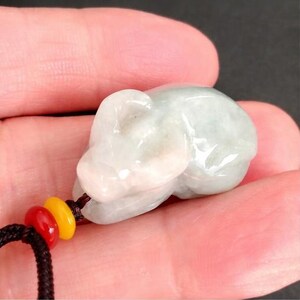 May include: A light green jade pendant carved in the shape of a small animal, possibly a mouse or a rat. The pendant is attached to a black cord with red and yellow beads. The jade has a translucent quality.