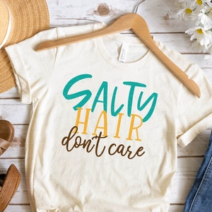 K&ouml;nnte beinhalten: Ein wei&szlig;es T-Shirt mit dem Text "Salty Hair don't care" in t&uuml;rkisfarbener und gelber Schrift.