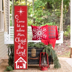May include: A red wooden sign with white lettering that reads "O Come let us adore Him, Christ the Lord". The sign has a star at the top and a manger scene at the bottom. The sign is hanging on a white porch swing with red and white plaid cushions.