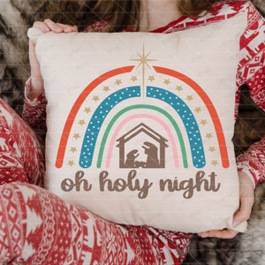 Może przedstawiać: Biała poduszka z kolorowym wzorem tęczy i brązową sylwetką sceny narodzin w środku. Tekst "oh holy night" jest nadrukowany pod tęczą.