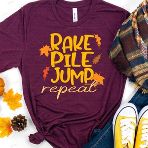 Peut inclure: Un t-shirt marron avec le texte "RAKE PILE JUMP repeat" en jaune avec des accents de feuilles orange.