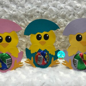 May include: Three yellow chick papercraft holders with pink, teal, and purple eggshells. Each chick is holding a chocolate egg candy.