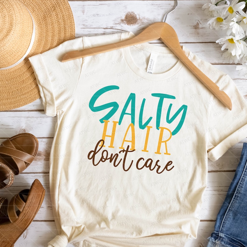 K&ouml;nnte beinhalten: Ein wei&szlig;es T-Shirt mit dem Text "Salty Hair don't care" in t&uuml;rkisfarbener und gelber Schrift.