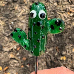 May include: A small, green glass cactus figurine featuring two side arms, black polka dot accents, and two white and black googly eyes. The cactus is attached to a thin, metallic rod extending from the bottom, creating a decorative garden stake.