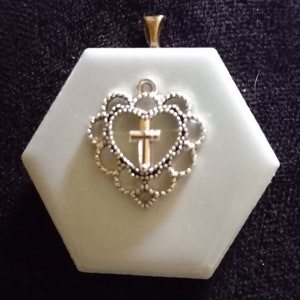 May include: A silver pendant with a heart-shaped design featuring a cross inside. The pendant is on a light blue hexagonal background.