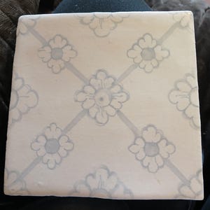 May include: Square, off-white tile with a repeating floral pattern in light blue. The flowers are arranged in a grid, with diagonal lines connecting them. The tile is likely ceramic or porcelain.