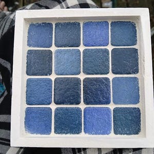 May include: A square, white-framed tray filled with sixteen blue square tiles. The tiles vary in shades from light to dark blue, creating a textured, mosaic-like appearance. The tray is resting on a black and white plaid surface.