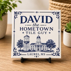 May include: A square white ceramic tile featuring dark blue decorative borders and text that reads DAVID the HOMETOWN TILE GUY. Below the text is a detailed blue illustration of a water tower labeled LAUREL MS and a building with a clock tower.