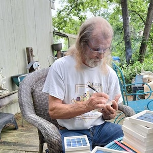 May include: A man seated outdoors, working on mosaic tiles. He is wearing a white t-shirt and blue jeans. Several mosaic tile squares are stacked nearby. The tiles are blue and white, set in a white frame.