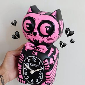 Puede incluir: Reloj de gato esqueleto rosa y negro con cola oscilante. La esfera del reloj es negra con números blancos y las palabras "Kit Cat" en blanco. El reloj está decorado con corazones y huesos rosas.