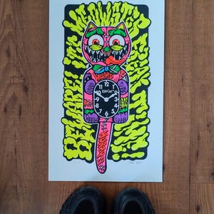 May include: A vibrant cat-shaped clock print featuring a pink cat face with a protruding tongue. The clock face is black, and the tail is pink. The print has a bright yellow and green background. The word "WEIRD" is above the clock.