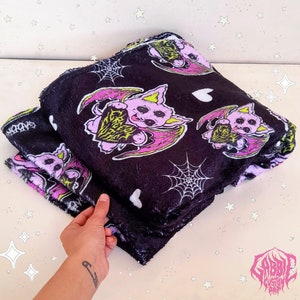 May include: A folded black blanket with a repeating pattern of a pink bat holding a heart, white hearts, and spiderwebs. The blanket has a soft, plush texture and the word "Sabbath" is printed on the edge.