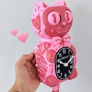 May include: Pink cat-shaped clock with a black face and white hands. The clock has a pink pendulum in the shape of a cat's tail with a heart and devil's tail. The clock face has the words "Kit Cat" and "LOVE HATE" printed on it.