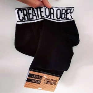 May include: A pair of black ankle socks with a white band at the top that reads "CREATE OR OBEY". The socks are folded and the brand label is visible at the bottom.