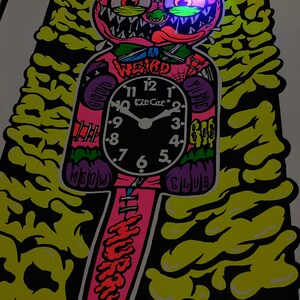 K&ouml;nnte beinhalten: Ein farbenfrohes, cartoonhaftes Uhrdesign mit einem Katzenkopf und dem Wort "WEIRD" &uuml;ber dem Zifferblatt. Das Zifferblatt zeigt "Kit Cat" und die Uhrzeit ist ungef&auml;hr 7:00 Uhr. Das Design enth&auml;lt die W&ouml;rter "MEOW CLUB" und "HURRY".