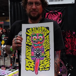 K&ouml;nnte beinhalten: Ein farbenfrohes Kunstwerk mit einem Cartoon-Katzen-Uhr-Design mit den Worten "WEIRD TIME" und "GOOD HORROR". Die Katzen-Uhr ist rosa, mit gelben und gr&uuml;nen Akzenten. Das Kunstwerk wird von einer Person auf einer Kunstmesse gehalten.