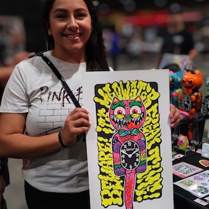 K&ouml;nnte beinhalten: Ein farbenfroher Kunstdruck mit einem Cartoon-Katzen-Uhr-Design. Die Katze hat einen rosa K&ouml;rper, gr&uuml;ne Augen und ein Ziffernblatt. Der Hintergrund ist gelb mit schwarzen Umrissen. Das Wort "WEIRD" steht &uuml;ber dem Ziffernblatt. Der Druck wird von einer Person gehalten.