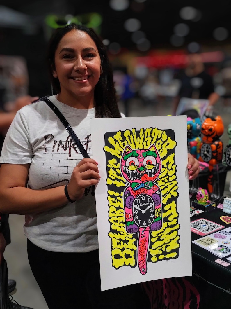 K&ouml;nnte beinhalten: Ein farbenfroher Kunstdruck mit einem Cartoon-Katzen-Uhr-Design. Die Katze hat einen rosa K&ouml;rper, gr&uuml;ne Augen und ein Ziffernblatt. Der Hintergrund ist gelb mit schwarzen Umrissen. Das Wort "WEIRD" steht &uuml;ber dem Ziffernblatt. Der Druck wird von einer Person gehalten.