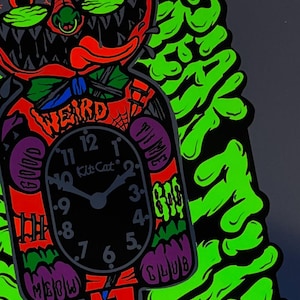 K&ouml;nnte beinhalten: Eine farbenfrohe Wanduhr im Cartoon-Stil mit einem schwarzen Zifferblatt und der Aufschrift "Kit Cat". Die Uhr hat einen rot-orangenen K&ouml;rper mit dem Wort "WEIRD" darauf. Das Design enth&auml;lt gr&uuml;ne, violette und schwarze Akzente.