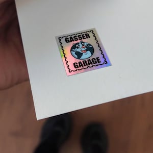 K&ouml;nnte beinhalten: Ein holografischer Aufkleber mit dem Text "GASSER GARAGE" und einem Cartoon-Globus mit einem l&auml;chelnden Gesicht. Der Aufkleber hat einen schwarzen, gezackten Rand und befindet sich auf einer wei&szlig;en Oberfl&auml;che.