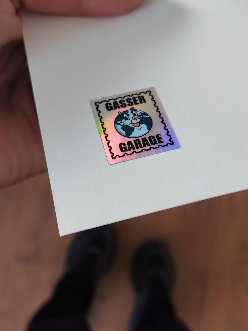 K&ouml;nnte beinhalten: Ein holografischer Aufkleber mit dem Text "GASSER GARAGE" und einem Cartoon-Globus mit einem l&auml;chelnden Gesicht. Der Aufkleber hat einen schwarzen, gezackten Rand und befindet sich auf einer wei&szlig;en Oberfl&auml;che.