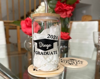 May include: A clear glass tumbler with a bamboo lid and a straw. The tumbler has a black and white sticker that says "2024 Diego GRADUATE". The tumbler is sitting on a wooden coaster.