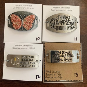 May include: Four metal connector charms with different designs. The first charm features a pink butterfly, the second charm has the text "Think happy thoughts", the third charm has the text "You are exactly where you need to be", and the fourth charm has the text "And though she be but little, she is fierce."