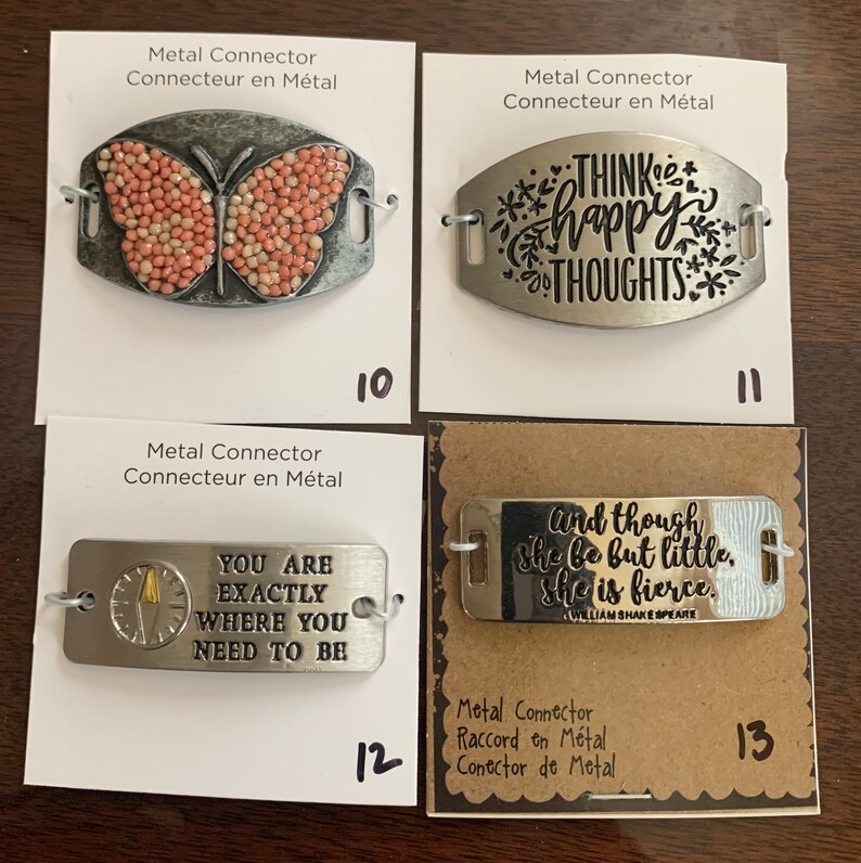 May include: Four metal connector charms with different designs. The first charm features a pink butterfly, the second charm has the text "Think happy thoughts", the third charm has the text "You are exactly where you need to be", and the fourth charm has the text "And though she be but little, she is fierce."