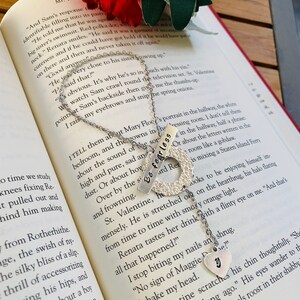 May include: Two silver necklaces with charms that say "Be Fearless" and "Y". The necklaces are on a book with a red and white floral design. The book has text about a character named Sam and Mary Floce.