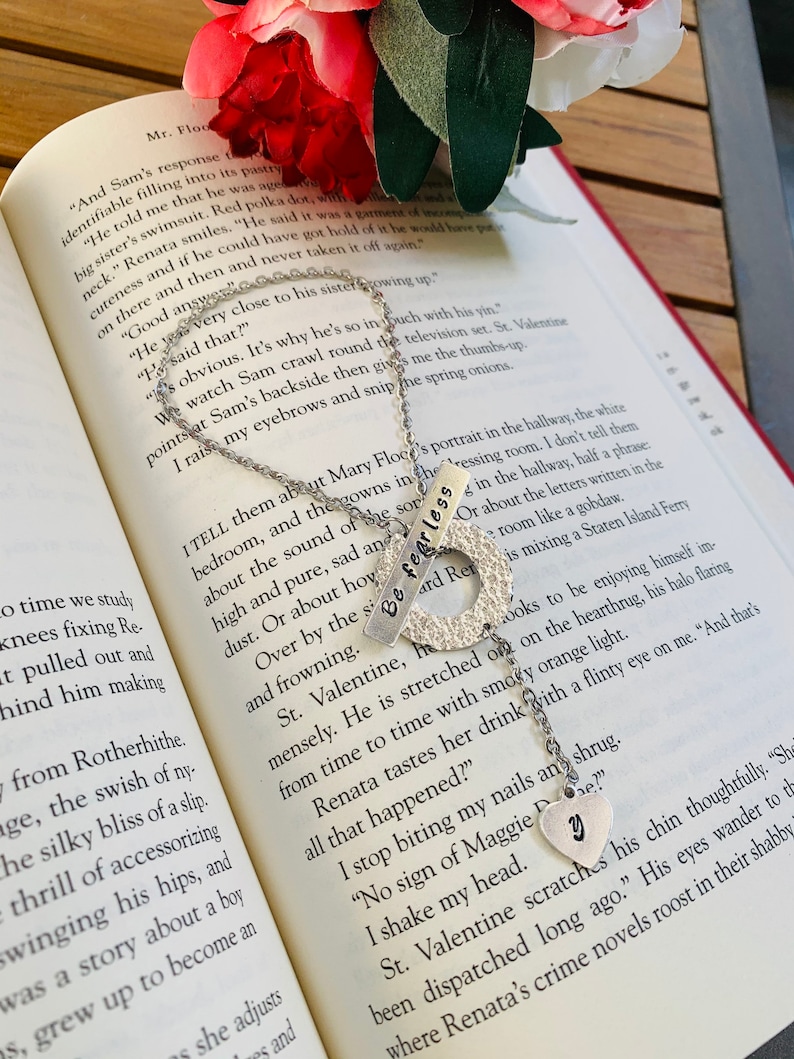 May include: Two silver necklaces with charms that say "Be Fearless" and "Y". The necklaces are on a book with a red and white floral design. The book has text about a character named Sam and Mary Floce.