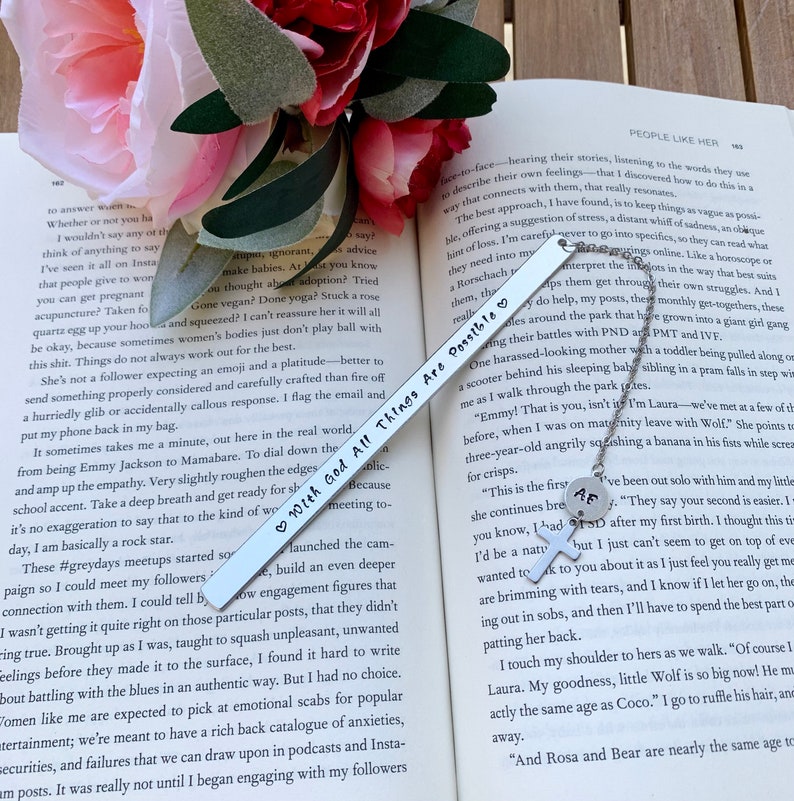 Pu&ograve; includere: Un segnalibro d'argento con un cuore e le parole "With God All Things Are Possible" incise su di esso. Il segnalibro &egrave; appoggiato su un libro con un fiore rosa sullo sfondo.
