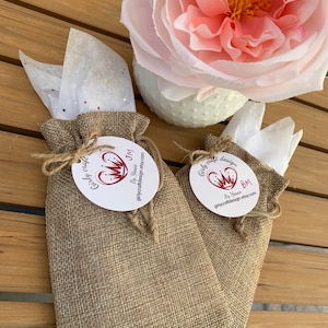 Pu&ograve; includere: Due piccoli sacchetti di juta con un'etichetta circolare bianca attaccata a ciascuno. Le etichette hanno un cuore rosso con una corona e il testo "Girly craft design" e "By Yassie" su di esse. I sacchetti sono su una superficie di legno.