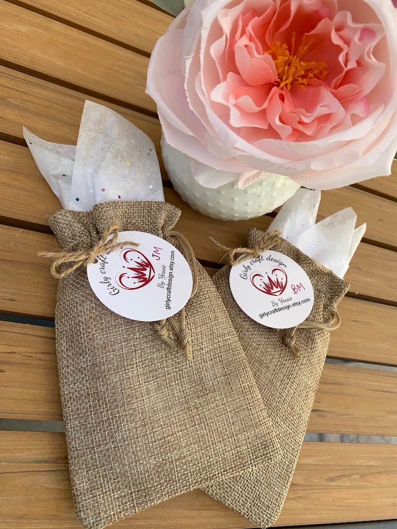 Pu&ograve; includere: Due piccoli sacchetti di juta con un'etichetta circolare bianca attaccata a ciascuno. Le etichette hanno un cuore rosso con una corona e il testo "Girly craft design" e "By Yassie" su di esse. I sacchetti sono su una superficie di legno.