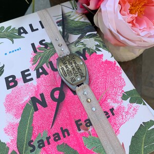 May include: A book titled "Ali is Beauty Now" by Sarah Fae, with a floral design cover. A bookmark with a metal charm that reads "Live in the sunshine, swim in the sea, drink the wild air - Emerson" is attached to the book.