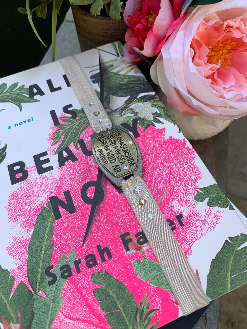 May include: A book titled "Ali is Beauty Now" by Sarah Fae, with a floral design cover. A bookmark with a metal charm that reads "Live in the sunshine, swim in the sea, drink the wild air - Emerson" is attached to the book.