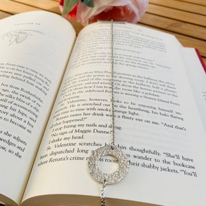 May include: A silver charm necklace with a round charm and a heart charm. The necklace is lying on a book with a red cover. The book is open to a page with text about a character named Renata.