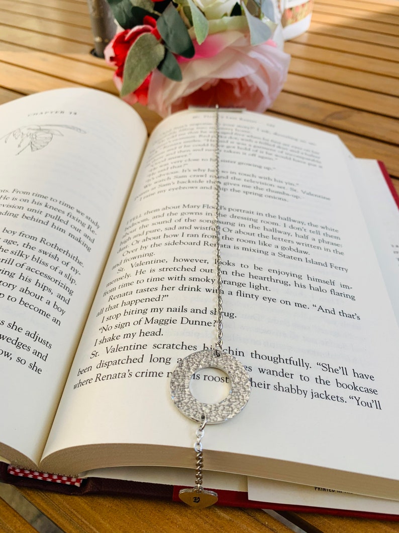 May include: A silver charm necklace with a round charm and a heart charm. The necklace is lying on a book with a red cover. The book is open to a page with text about a character named Renata.