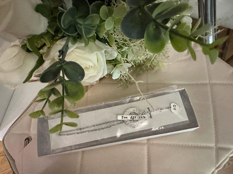 May include: A silver necklace with a charm that says "You got this" and a heart charm. The necklace is in a white box with a white background.