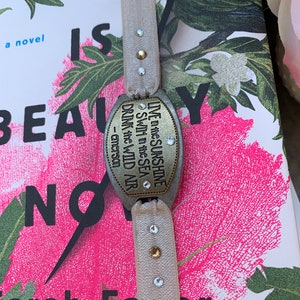 May include: A beige ribbon bookmark with a silver charm that reads "LIVE in the SUNSHINE SWIM in the SEA DRINK the WILD AIR - emerson". The charm has rhinestones on it. The bookmark is on top of a book with a floral design.