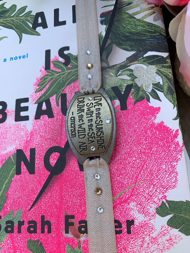 May include: A beige ribbon bookmark with a silver charm that reads "LIVE in the SUNSHINE SWIM in the SEA DRINK the WILD AIR - emerson". The charm has rhinestones on it. The bookmark is on top of a book with a floral design.