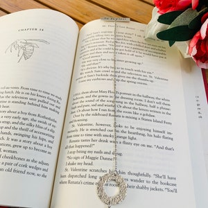 May include: A silver bookmark with a circular design featuring a cutout pattern. The bookmark is resting on an open book with text about a fictional character named St. Valentine.