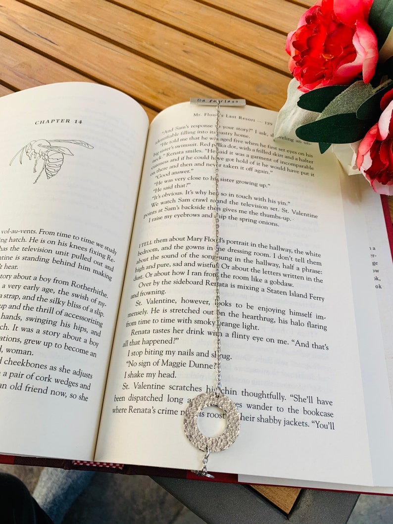 May include: A silver bookmark with a circular design featuring a cutout pattern. The bookmark is resting on an open book with text about a fictional character named St. Valentine.