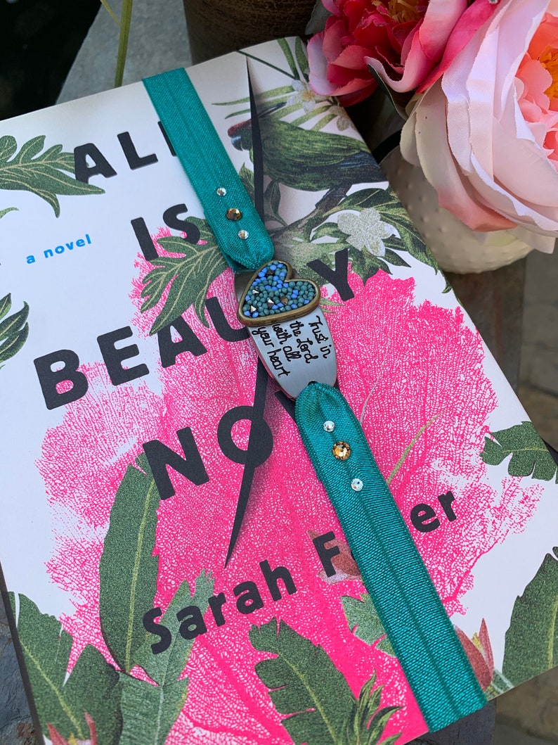 May include: A teal blue bookmark with rhinestones and a heart-shaped charm with the text "Trust in the Lord with all your heart". The bookmark is on a book with a pink and green floral design and the title "All is Beauty Now" by Sarah Fler.