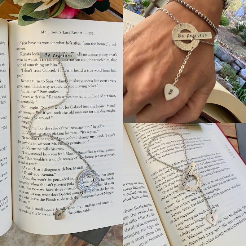 May include: A silver charm bracelet with a heart charm, a round charm with the words "Be fearless" engraved on it, and a chain with a heart charm. The bracelet is on a book with the text "Mr. Flood's Last Resort" visible.