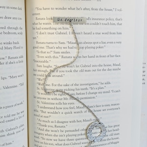 May include: A silver necklace with a heart charm and a round charm with the words "Be fearless" engraved on it. The necklace is lying on an open book with text about a mystery.