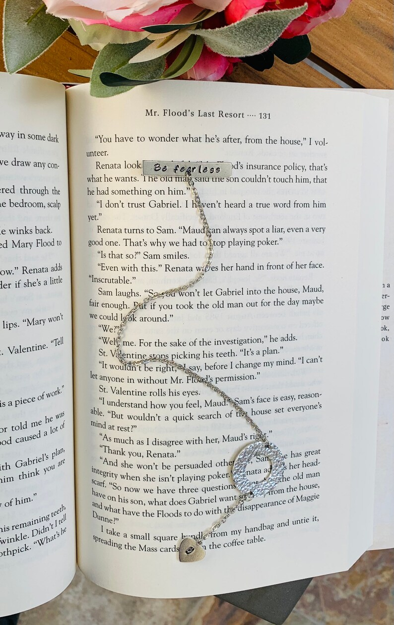 May include: A silver necklace with a heart charm and a round charm with the words "Be fearless" engraved on it. The necklace is lying on an open book with text about a mystery.