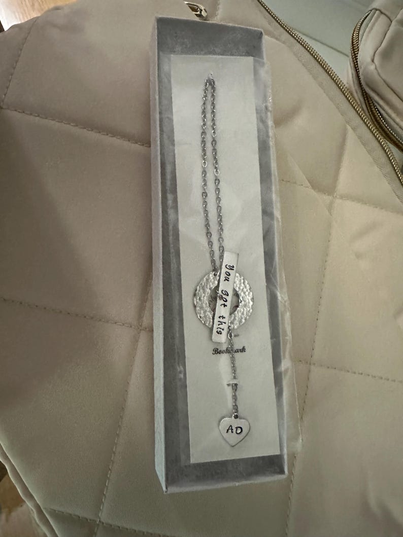 May include: Silver necklace with a heart charm that says "AD" and a round charm with the text "You got this" hanging from a silver chain. The necklace is in a white box.