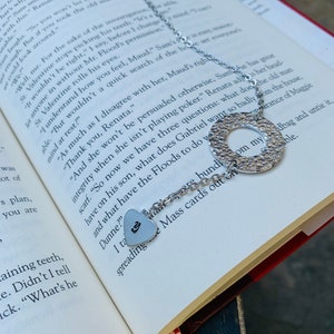 May include: A silver necklace with a heart charm and a round charm with a textured surface. The necklace is lying on a book with text about a fictional character named Maud.